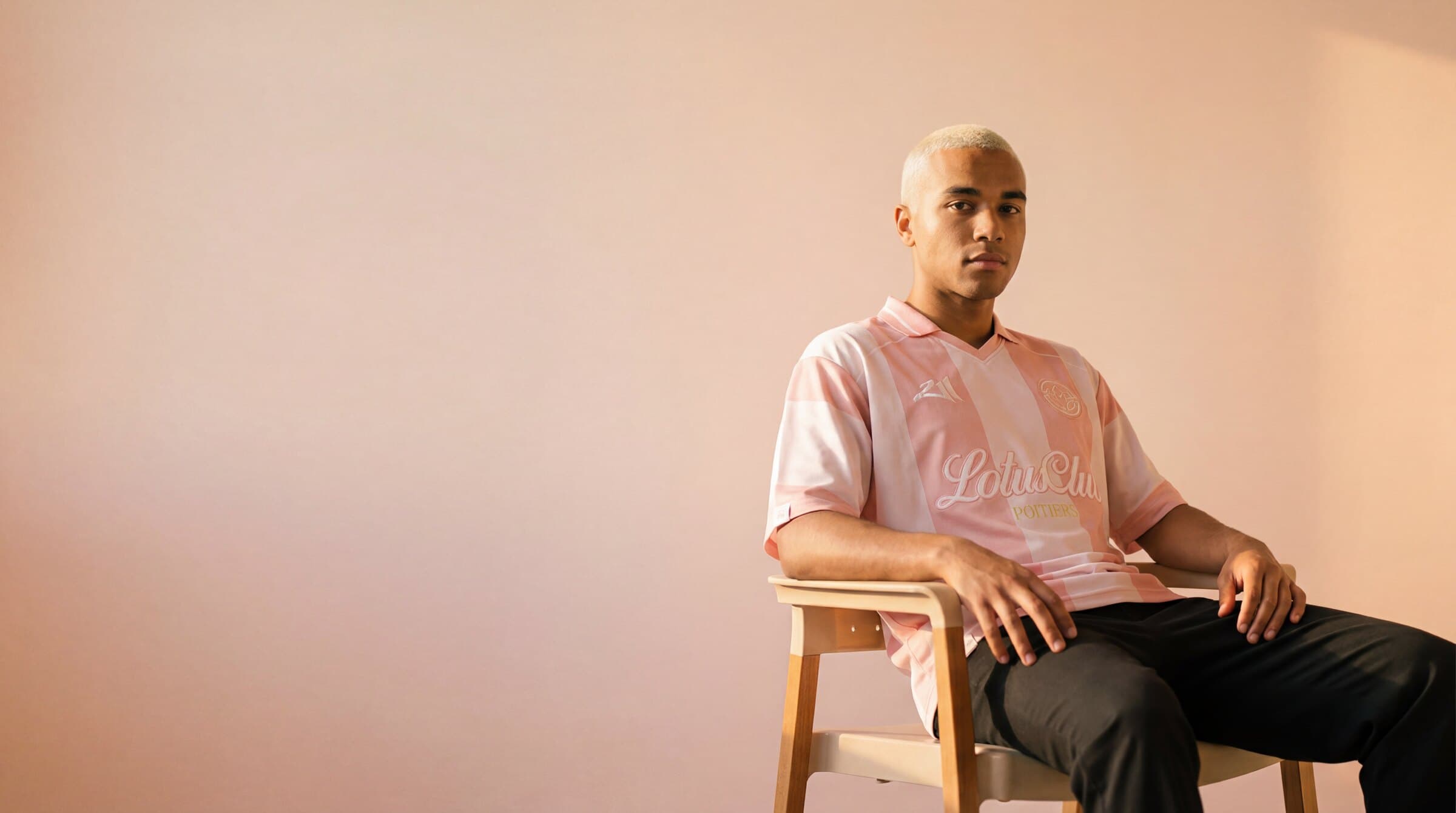 Mannequin portant le polo rose Lotus Club Clothing, assis sur une chaise devant un mur pastel